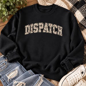 Peut inclure: Un sweat-shirt noir à col rond avec le mot DISPATCH imprimé dans une grande police universitaire arquée. Les lettres présentent un motif léopard beige et marron avec un fin contour blanc. Le sweat-shirt a des manches longues et un col côtelé.
