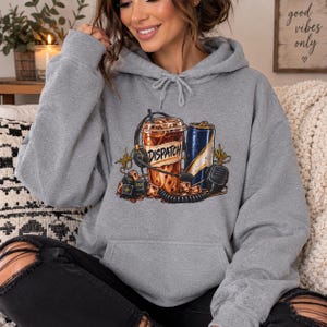 May include: A heather grey hooded sweatshirt featuring a graphic print of an iced coffee cup labelled DISPATCH, a blue energy drink can, a headset, a microphone, and computer keys. The hoodie has long sleeves, a front kangaroo pocket, and a drawstring.