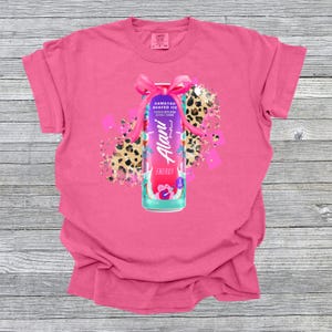 Puede incluir: Camiseta rosa con un gr&aacute;fico de una lata de bebida energ&eacute;tica Alani Nu, adornada con un lazo rosa, sobre un fondo de estampado de leopardo y confeti rosa. La lata es turquesa, morada y rosa, con "Hawaiian Shaved Ice" y "Energy".