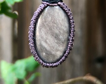 May include: A handcrafted pendant necklace featuring a polished, oval-shaped stone in shades of gray and silver. The stone is encased in a macrame-style weave of dark purple cord, suspended from a black woven cord necklace.