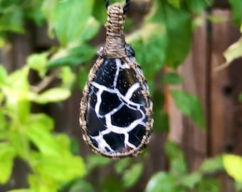 May include: A black and white agate pendant with a brown macrame wrap, hanging from a black cord.