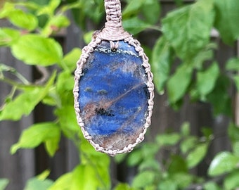Puede incluir: Un colgante de piedra azul y marrón con un diseño de nudo macramé, colgado de un cordón negro.
