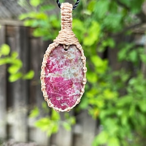 May include: A pink and white gemstone pendant with a knotted cord wrap, hanging from a black cord.
