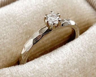 May include: A delicate diamond engagement ring with a textured band, displayed in a beige velvet ring box.