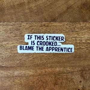May include: A white sticker with the text "IF THIS STICKER IS CROOKED... BLAME THE APPRENTICE" in a dark blue font. The sticker is placed on a wooden surface with a natural wood grain.
