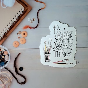 May include: An overhead shot of craft supplies on a light wood surface. Includes a notebook, orange thread, buttons, beads, yarn, and a sticker that reads "I just want to drink coffee & make pretty things."