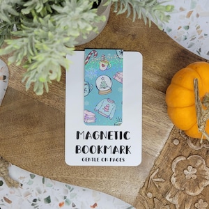 May include: A white magnetic bookmark with a teal patterned design featuring a candy cane, coffee cup, snow globe, and sweater. The text "MAGNETIC BOOKMARK GENTLE ON PAGES" is printed in black. The bookmark is on a wooden surface with a small pumpkin.