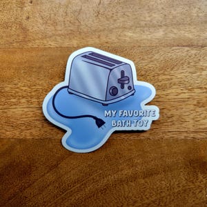 May include: A sticker of a silver toaster with a blue cord and a blue puddle around it. The text "MY FAVORITE BATH TOY" is printed below the toaster.