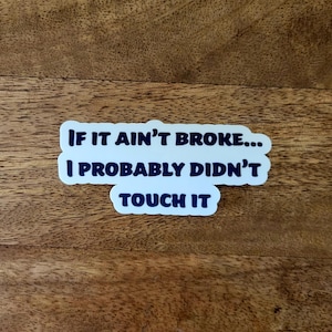 May include: A white sticker with the text "IF IT AIN'T BROKE... I PROBABLY DIDN'T TOUCH IT" in dark blue lettering. The sticker is on a wooden surface with a natural wood grain.