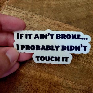 May include: White sticker with the text "IF IT AIN'T BROKE... I PROBABLY DIDN'T TOUCH IT" in black, sans-serif font. The sticker has a white border and is held against a wooden surface.