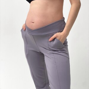May include: Gray maternity pants with a crossover waistband and pockets. The pants have a tailored fit and are made of a soft, stretchy fabric. The design includes vertical seams down the front of the legs.