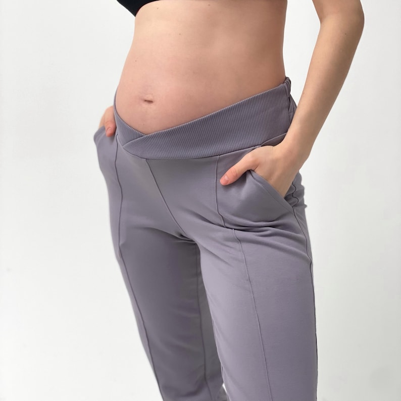 May include: Gray maternity pants with a crossover waistband and pockets. The pants have a tailored fit and are made of a soft, stretchy fabric. The design includes vertical seams down the front of the legs.