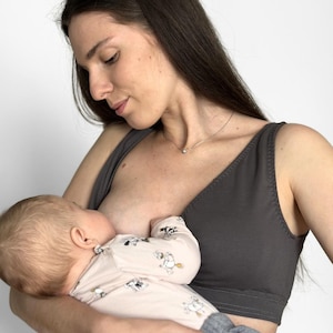May include: A dark charcoal gray nursing bra featuring a V-neckline, wide shoulder straps, and a supportive under-bust band. The garment is constructed with visible stitching along the neckline and hem, providing a functional design for breastfeeding.
