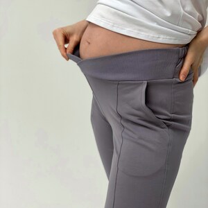 May include: Gray maternity pants with a wide, ribbed waistband and a relaxed fit. The pants have a pocket on the side and are made of a soft, stretchy material. The pants are designed to accommodate a growing belly.