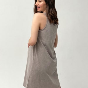 May include: A light brown, sleeveless dress with a scoop neckline. The dress is knee-length and made of a soft, jersey-like material. The model is smiling and looking over their shoulder.