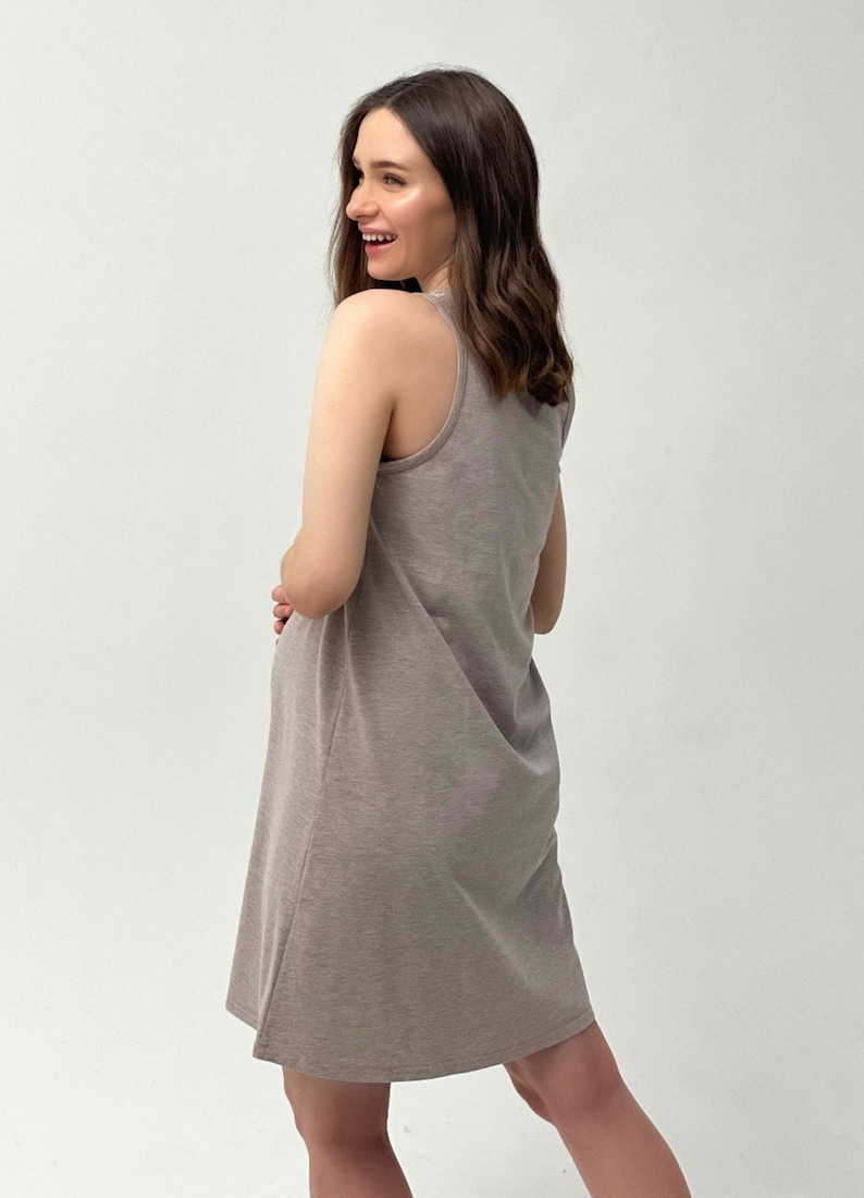 May include: A light brown, sleeveless dress with a scoop neckline. The dress is knee-length and made of a soft, jersey-like material. The model is smiling and looking over their shoulder.