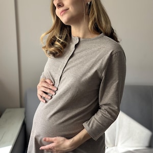 May include: A light brown maternity dress with long sleeves and a button-up front. The dress is made of a soft fabric and is designed for pregnancy. The woman is holding her baby bump.