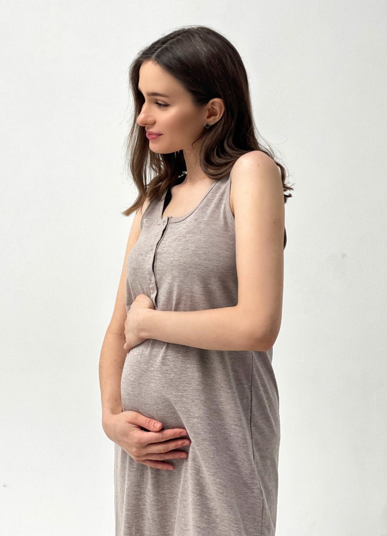 May include: A sleeveless, button-down, taupe-colored maternity dress. The dress is made of a soft, flowing fabric and is designed to accommodate a growing belly. The model is wearing the dress and holding her belly.