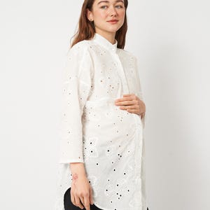 May include: White eyelet maternity shirt with a mandarin collar and button-down front. The long-sleeved shirt features floral cut-out details and a slightly longer hemline in the front. The shirt is paired with black pants.