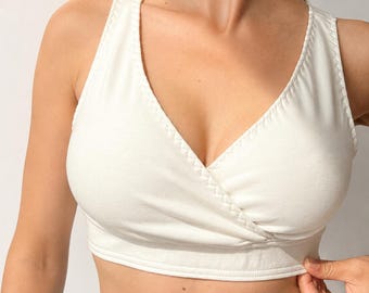 May include: A white V-neck crop top featuring a wrap-style front design with thick shoulder straps and a wide hem band. The garment is constructed with visible stitched detailing along the neckline and armholes, providing a clean finish.