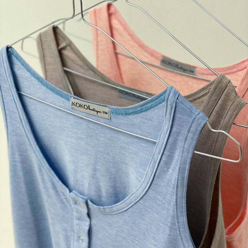 May include: Three tank tops in pastel colors hang on metal hangers. The front tank top is light blue with a button-up front and a blue trim. The other two tank tops are a taupe and a light pink. Each tank top has a tag that reads "KOKO boutique S/M".