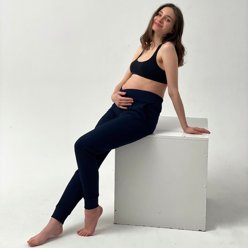 May include: A black sports bra and navy blue sweatpants. The sweatpants have a high waistband and a relaxed fit. The model is sitting on a white cube. The background is plain white.