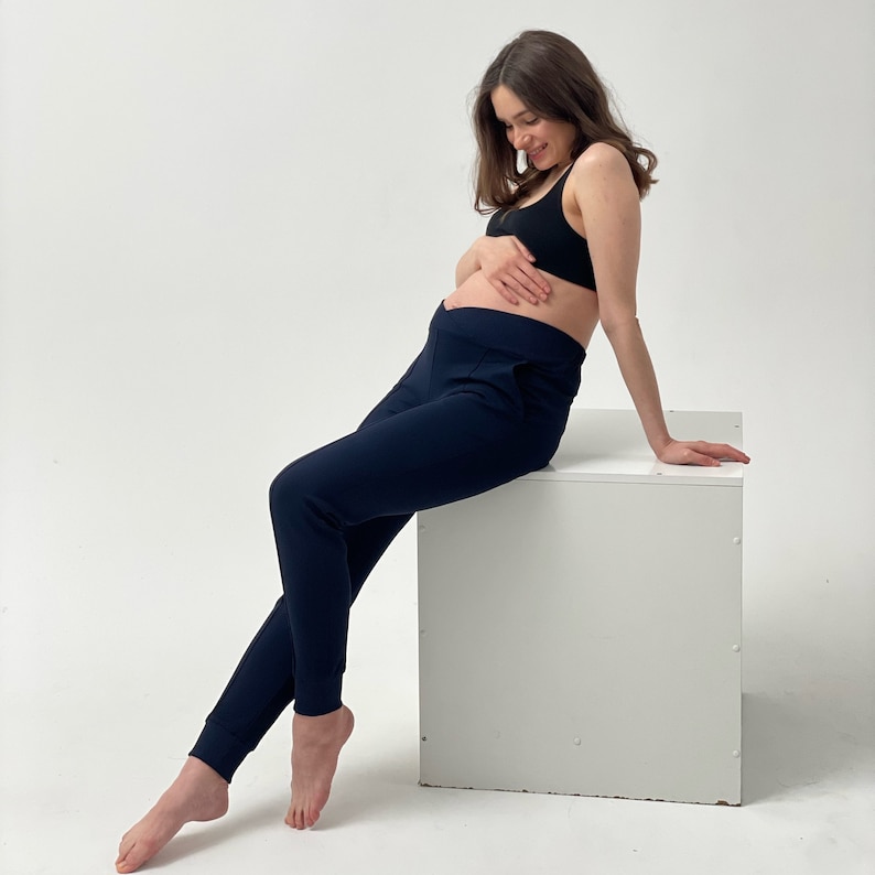 May include: Navy blue maternity joggers with a high waistband and cuffed ankles. The joggers are paired with a black sports bra. The model is sitting on a white cube against a white background.