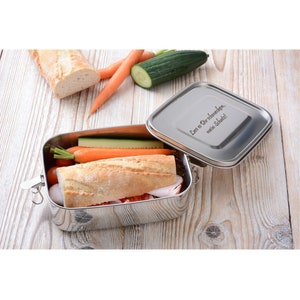 Peut inclure: Une boîte à lunch en acier inoxydable contenant un sandwich, des carottes et un concombre. Le couvercle porte une inscription qui dit "Lass es dir schmecken, mein Schatz!"