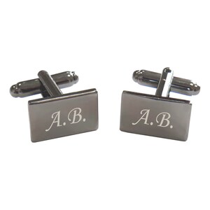 Könnte beinhalten: Ein Paar schwarze Manschettenknöpfe mit rechteckigem Design. Die Manschettenknöpfe sind mit den Initialen "A.B." graviert.