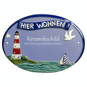 Può includere: Un cartello per la casa in ceramica blu e bianca con un faro, una barca a vela e una colomba. Il cartello recita "Hier Wohnen" e "Keramikschild mit Deiner persönlichen Gravur".