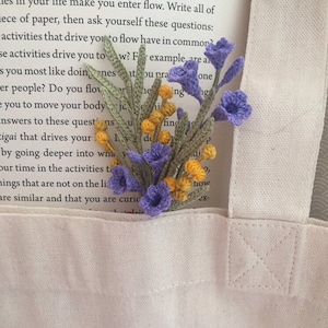 Op de afbeelding: Een close-up van een gehaakt bloemenboeket met paarse en gele bloemen. Het boeket ligt op een witte canvas tas met een boek zichtbaar op de achtergrond.