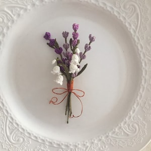 May include: A small bouquet of crocheted flowers in shades of purple and white, tied together with a brown ribbon. The flowers are arranged on a white plate with a decorative border.