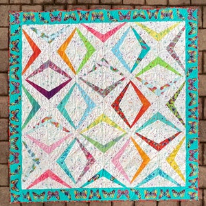 Könnte beinhalten: Ein farbenfroher Quilt mit einem geometrischen Design. Der Quilt hat einen weißen Hintergrund mit rautenförmigen Formen in verschiedenen Farben, darunter Orange, Rosa, Grün und Blau. Ein türkisfarbener Rand mit Schmetterlingsmustern umrahmt den Quilt.
