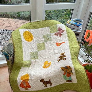 Peut inclure: Une courtepointe faite à la main avec un fond blanc et une bordure verte. La courtepointe présente des images appliquées d'un soleil, d'un papillon, d'un oiseau, d'un ours en peluche, d'un canard, d'un chien et d'un enfant. La courtepointe est drapée sur une chaise à motifs.