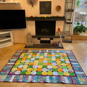 Könnte beinhalten: Ein farbenfroher Patchwork-Decken mit einem geometrischen Muster aus Quadraten und Kreisen in Grün-, Gelb-, Orange- und Lilatönen. Die Decke liegt auf einem Holzboden.