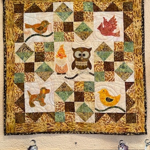 Könnte beinhalten: Ein gesteppter Wandbehang mit Patchwork-Muster, das einen braunen und weißen Hund, einen gelben Vogel, einen roten Vogel, eine braune und weiße Eule und einen Zwerg zeigt. Der Hintergrund ist eine Mischung aus braunen, grünen und weißen Stoffen.