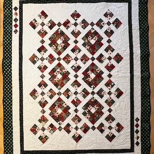 Op de afbeelding: Een handgemaakte quilt met een wit midden en een donkergroene rand. De quilt heeft een herhalend patroon van rode, groene en bruine bloemen vierkanten gerangschikt in een ruitvormig ontwerp. De rand heeft een stippenpatroon.