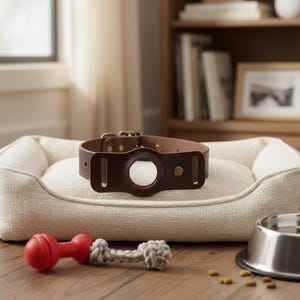Könnte beinhalten: Ein dunkelbraunes Haustierhalsband aus Leder mit einer kreisförmigen Aussparung für ein Ortungsgerät. Das Halsband verfügt über messingfarbene Metallbeschläge, darunter eine Schnalle und eine Niete, und liegt auf einem cremefarbenen Haustierbett.