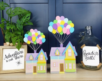 Könnte beinhalten: Zwei bunte Papierhäuser mit violetten Dächern und gelben Wänden werden von Bündeln bunter Ballons in die Luft gehoben. Die Häuser sind mit Fenstern und Türen verziert. Das kleinere Haus hat ein Schild mit der Aufschrift "Abenteuer ist da draußen". Das größere Haus hat ein Schild mit der Aufschrift "Paradise Falls".