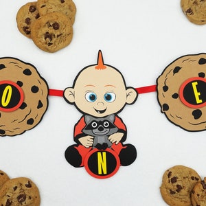 Può includere: Uno striscione rosso e nero con le lettere "ONE" in cerchi marroni su uno sfondo di biscotti marroni. La lettera centrale "N" è su un cerchio marrone con sfondo giallo. Un personaggio dei cartoni animati di un bambino con i capelli rossi e un vestito rosso e nero tiene un procione. Lo striscione è decorato con biscotti marroni.