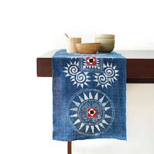 Puede incluir: Un camino de mesa azul con diseños blancos en forma de sol y acentos rojos y negros. El camino de mesa está sobre una mesa de madera con cuencos de madera y una cuchara de madera.