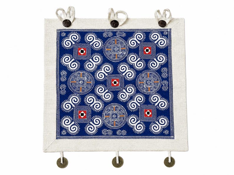 May include: A square textile wall hanging featuring a deep blue center panel with white spiral patterns, geometric squares, and circular motifs. The piece is framed by a light beige border and includes three top loops and three bottom hanging coins.