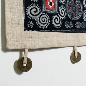 May include: A rectangular textile wall hanging featuring a black fabric panel with white spiral patterns and red square accents. The panel is bordered by a light beige linen-like fabric and includes three hanging bronze-colored metal coins.
