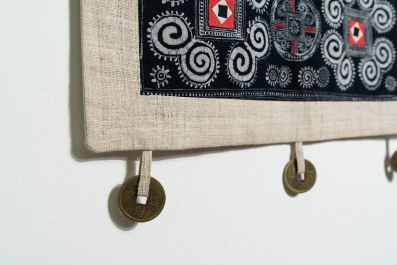 May include: A rectangular textile wall hanging featuring a black fabric panel with white spiral patterns and red square accents. The panel is bordered by a light beige linen-like fabric and includes three hanging bronze-colored metal coins.