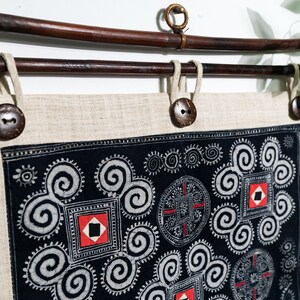 May include: A rectangular beige linen wall hanging suspended from a dark brown wooden rod. The fabric features a black panel with white spiral patterns, red square accents, and circular motifs. Three dark brown buttons secure the fabric to the rod.