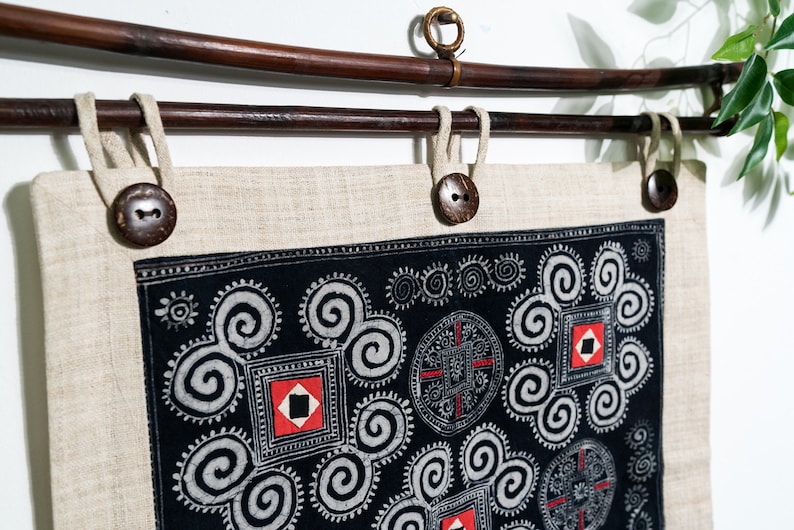 May include: A rectangular beige linen wall hanging suspended from a dark brown wooden rod. The fabric features a black panel with white spiral patterns, red square accents, and circular motifs. Three dark brown buttons secure the fabric to the rod.