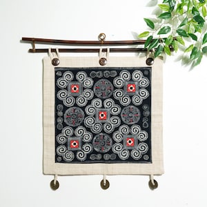 May include: A square wall hanging featuring a black fabric panel with white spiral patterns and red and white geometric squares. The panel is mounted on a dark brown bamboo rod with three dark buttons at the top and three metal coins at the bottom.