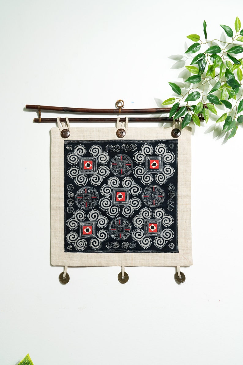 May include: A square wall hanging featuring a black fabric panel with white spiral patterns and red and white geometric squares. The panel is mounted on a dark brown bamboo rod with three dark buttons at the top and three metal coins at the bottom.