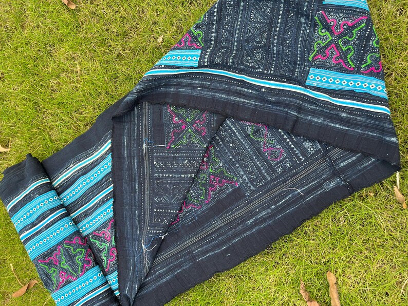 May include: A dark blue indigo fabric with intricate white and blue embroidery. The fabric has a geometric pattern with a repeating design of small squares and diamonds. The fabric is folded in half and the edges are frayed.