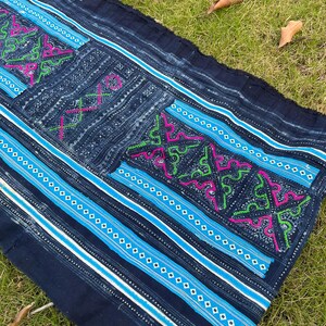 May include: A dark blue fabric with a white and blue striped pattern and intricate embroidery in pink, green, and white. The fabric has a frayed edge and is lying on a bed of green grass.